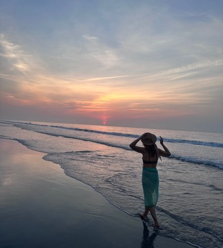 Person on beach at sunset, wearing hat.
